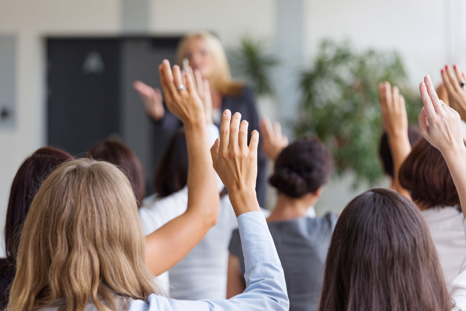 Der Blick aus dem Publikum einer Veranstaltung auf eine Moderatorin, die jungen Frauen im Publikum heben ihre Hände. | XXtalents - die Jobbörse für Frauen