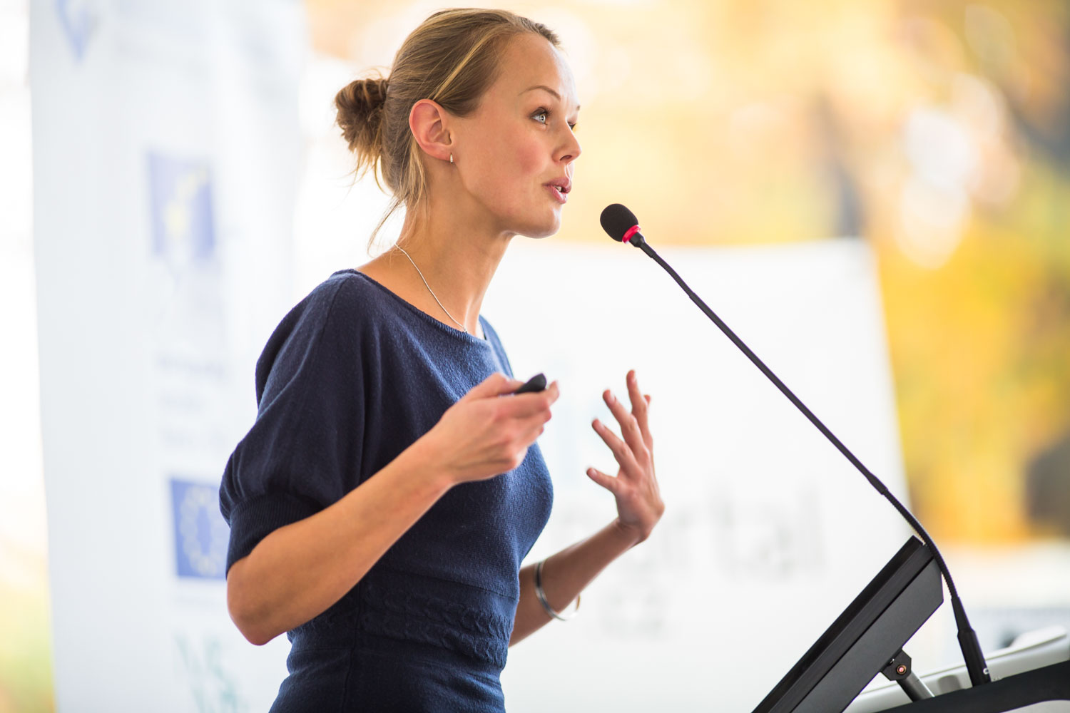 Eine junge Frau in blauem Pullover und mit blonden langen Haaren, die zu einem Knoten am Hinterkopf gebunden sind, spricht in ein Mikrofon an einem Rednerpult auf einer Veranstaltung. | XXtalents - die Jobbörse für Frauen